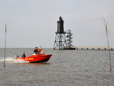 Ein rotes Motorboot der DLRG-Ortsgruppe Dorum fährt über das Wasser. Auf der Seite des Bootes steht gut sichtbar ‚DLRG‘. An Bord befinden sich mehrere Personen mit Rettungswesten. Im Hintergrund erhebt sich der Leuchtturm Obereversand. Im Vordergrund ragen zwei Pricken aus dem Wasser. 