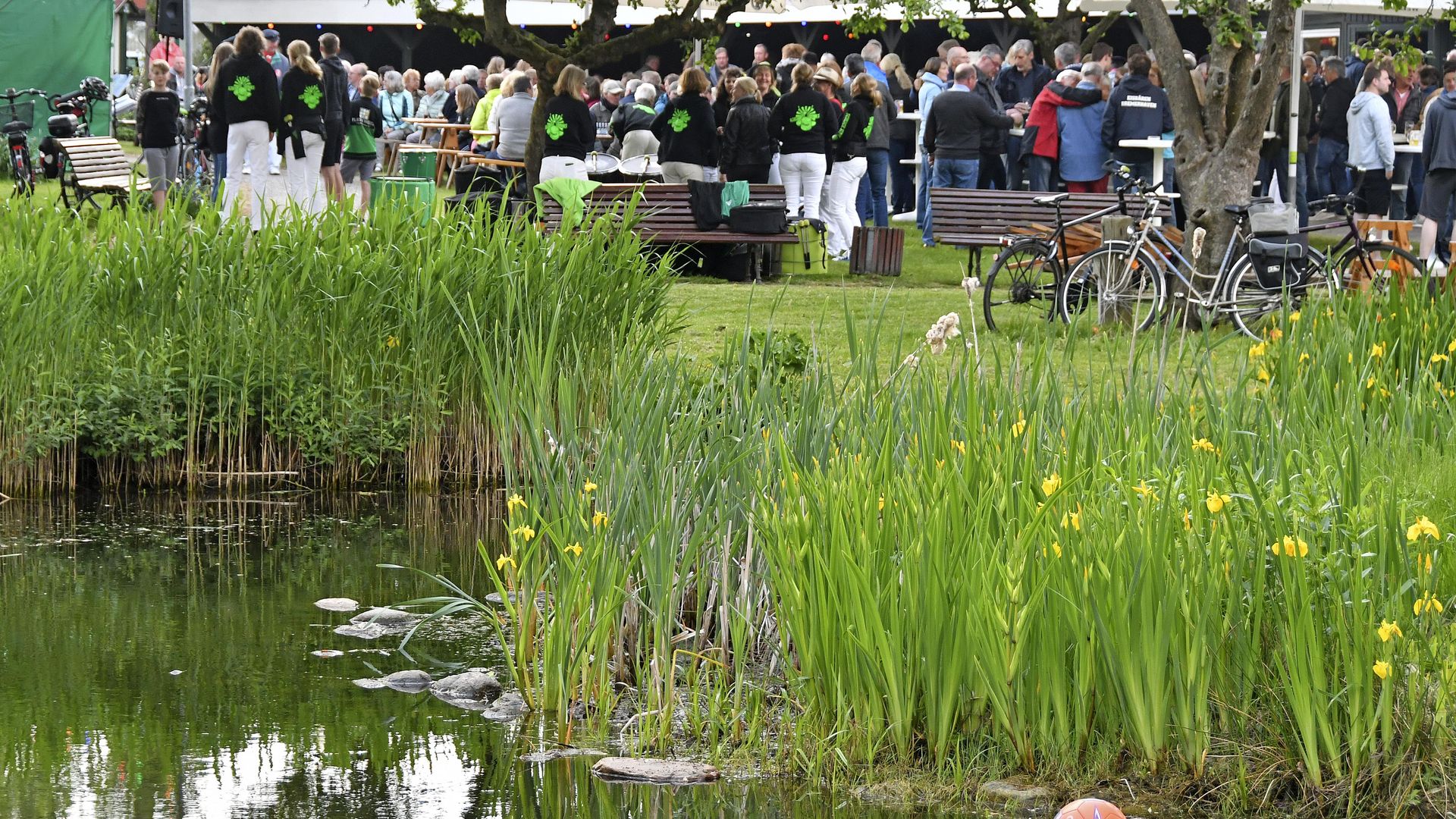Grille Wremen: Im Vordergrund liegt ein kleiner Teich mit grünen Schilfpflanzen und gelben Blumen am Ufer, auf der Wasseroberfläche schwimmt ein rot-blauer Ball. Im Hintergrund stehen zahlreiche Menschen in Gruppen auf einer Wiese unter Bäumen.
