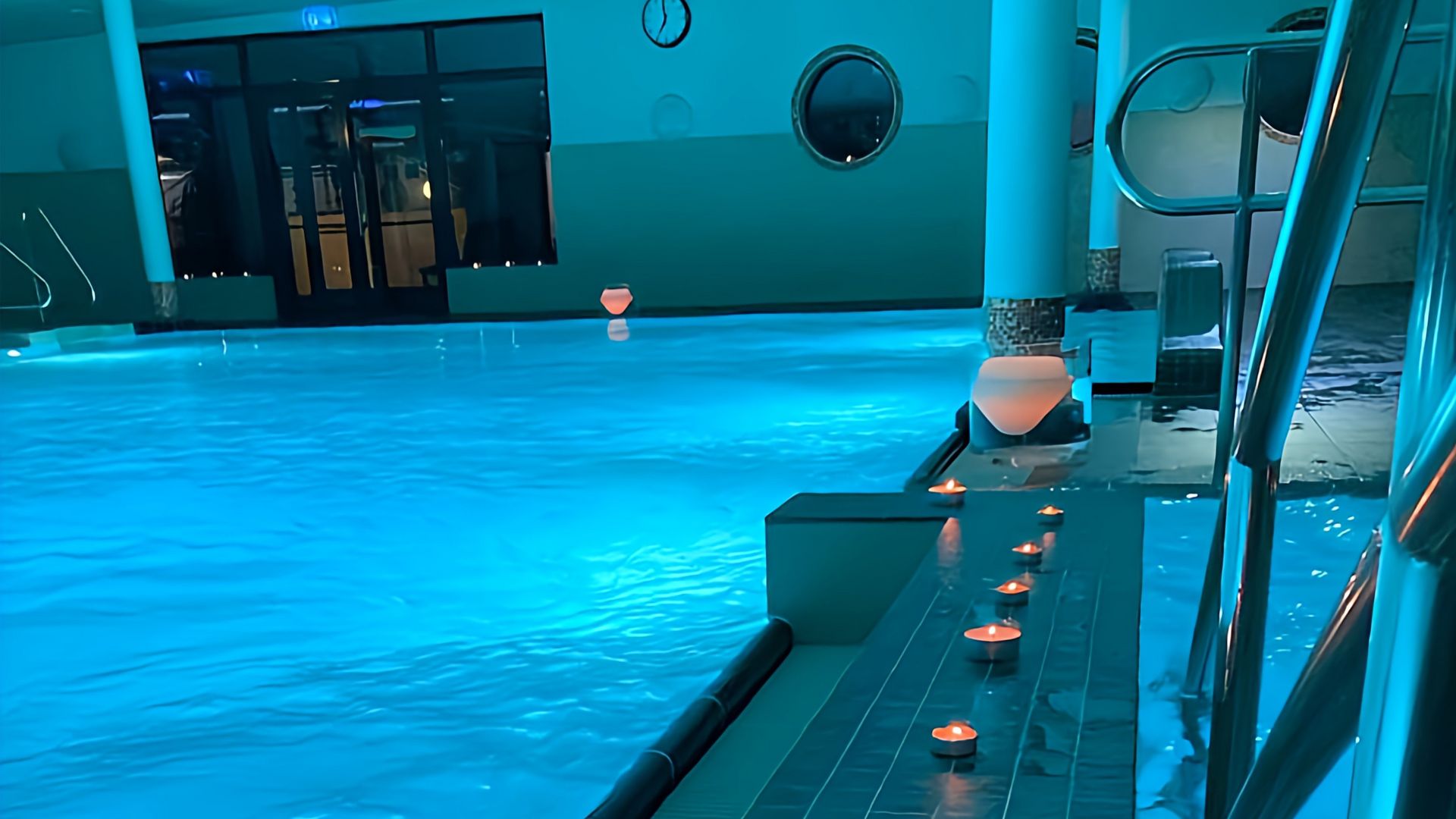 Lichterabend im Watt’n Bad: Das Hallenbad ist in sanftes blaues Licht getaucht, das eine ruhige und entspannende Stimmung erzeugt. Entlang des Beckenrandes stehen zahlreiche kleine Kerzen, deren warmes Licht sich im Wasser spiegelt. 