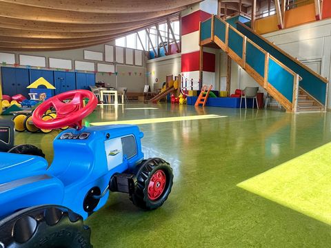 Innenansicht des Kinderspielhauses in Dorum-Neufeld: Der große, helle Raum ist mit grünem Bodenbelag ausgestattet und wird von Tageslicht durch mehrere Fenster erhellt. Im Vordergrund steht ein blaues Spielzeug-Traktor mit rotem Lenkrad. Im Hintergrund führt eine Holztreppe zu einer oberen Spielebene mit bunten Seitenwänden. Über den Raum verteilt liegen verschiedene Spielgeräte wie Bälle und kleine Rutschen. 