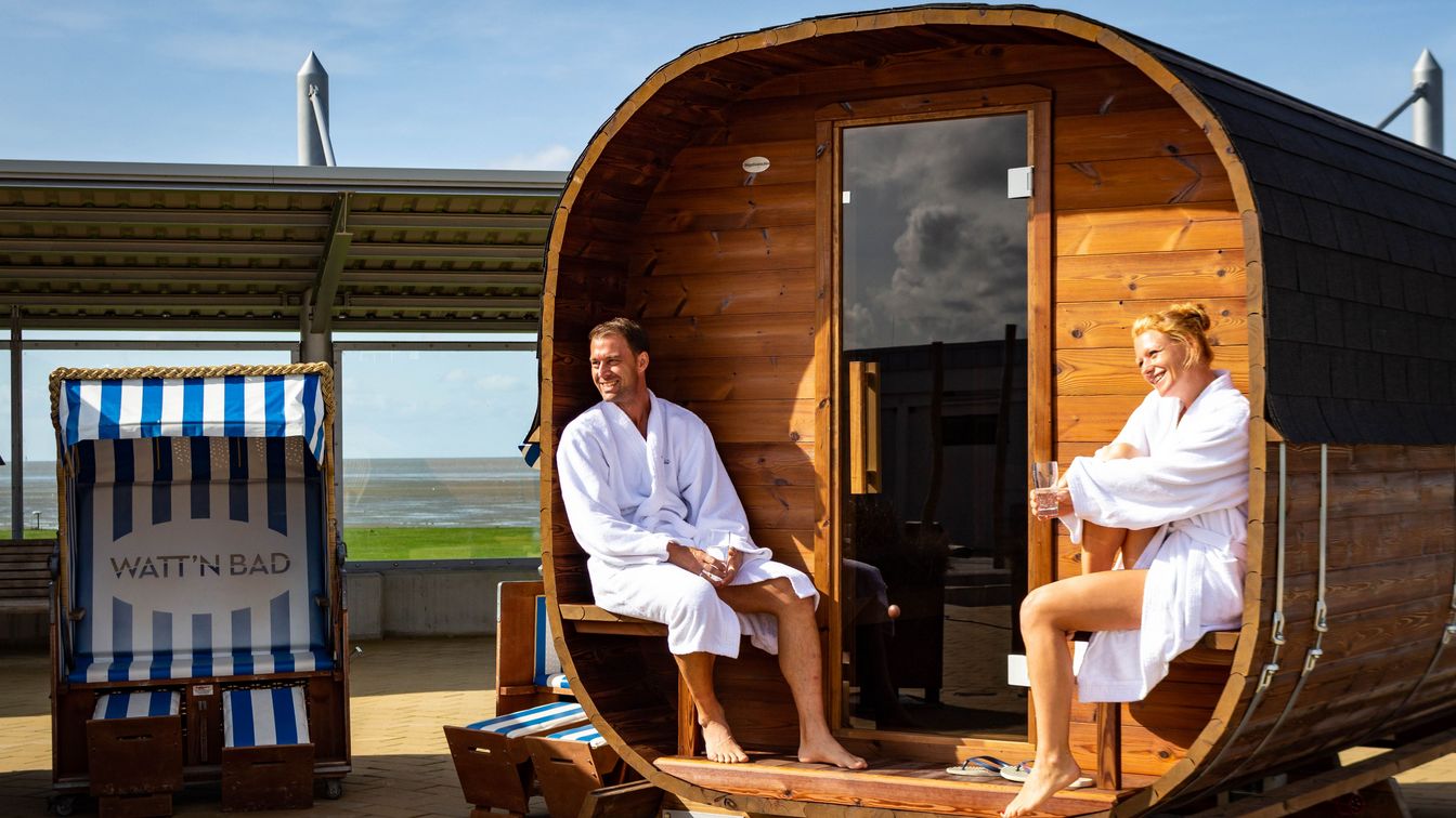 Eine Frau und ein Mann befinden sich vor der Fass-Sauna im Watt´n Bad, im weißen Bademantel und genießen den Sonnenschein. Im Hintergrund sieht man den Grünstrand und das Watt bei Ebbe.
