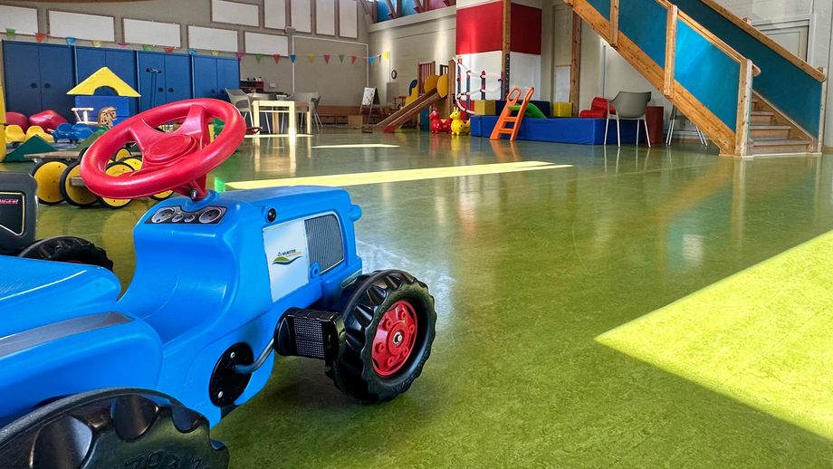 Innenansicht des Kinderspielhauses in Dorum-Neufeld: Der große, helle Raum ist mit grünem Bodenbelag ausgestattet und wird von Tageslicht durch mehrere Fenster erhellt. Im Vordergrund steht ein blaues Spielzeug-Traktor mit rotem Lenkrad. Im Hintergrund führt eine Holztreppe zu einer oberen Spielebene mit bunten Seitenwänden. Über den Raum verteilt liegen verschiedene Spielgeräte wie Bälle und kleine Rutschen. 
