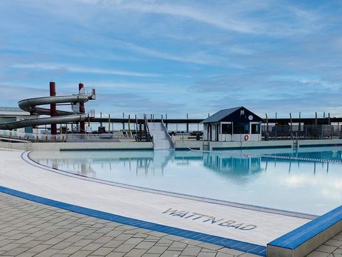 Der Wellenfreibadbereich im Watt’n Bad Dorum-Neufeld: Im Vordergrund liegt der Große Wellenfreibadbereich vom Watt'n Bad in Dorum-Neufeld mit klarem, blauem Wasser. Am weißen Beckenrand steht gut sichtbar der Schriftzug ‚WATT’N BAD‘. Im Hintergrund erhebt sich eine hohe Wasserrutsche mit mehreren Kurven, die für Spaß und Action sorgt. Der Himmel ist blau mit wenigen Wolken.