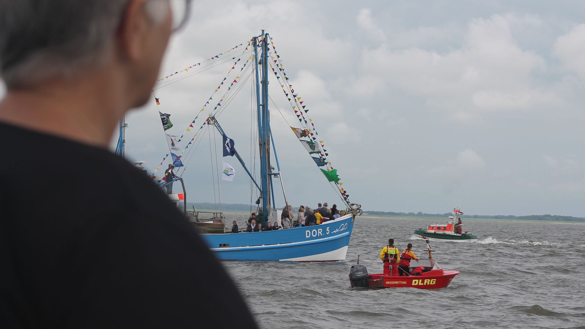 Beim Kutterkorso während der Dorumer Hafentage fährt ein blauer Fischkutter mit der Aufschrift ‚DOR 5‘ über das Wasser. Das Schiff ist mit bunten Flaggen und Wimpeln geschmückt und trägt mehrere Personen an Bord. Rechts daneben befindet sich ein rotes Rettungsboot der DLRG mit drei Personen in Rettungswesten sowie ein weiteres grün-weißes Boot im Hintergrund. Der Himmel ist bewölkt.