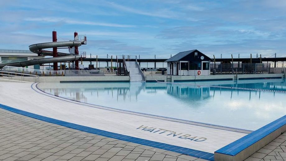 Der Wellenfreibadbereich im Watt’n Bad Dorum-Neufeld: Im Vordergrund liegt der Große Wellenfreibadbereich vom Watt'n Bad in Dorum-Neufeld mit klarem, blauem Wasser. Am weißen Beckenrand steht gut sichtbar der Schriftzug ‚WATT’N BAD‘. Im Hintergrund erhebt sich eine hohe Wasserrutsche mit mehreren Kurven, die für Spaß und Action sorgt. Der Himmel ist blau mit wenigen Wolken.