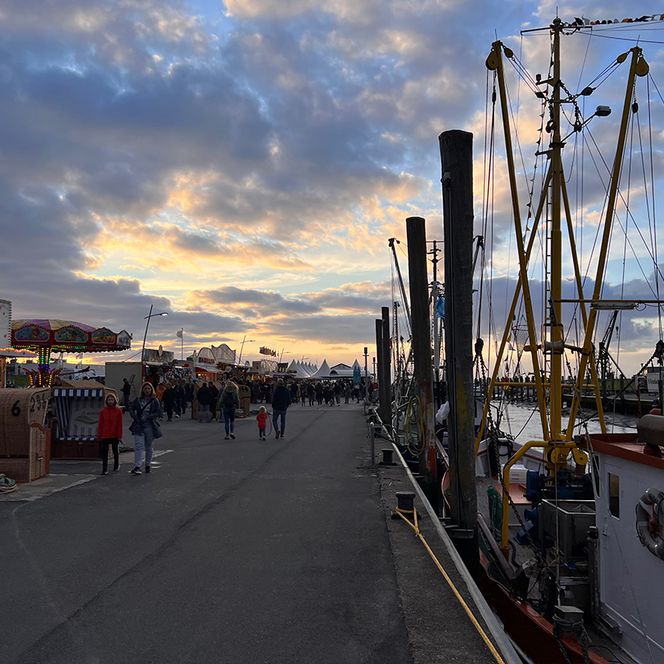 Abendstimmung auf der Hafenmeile bei den Dorumer Hafentagen: Links stehen beleuchtete Verkaufsstände mit bunten Markisen, vor denen Menschen entlangschlendern. Rechts liegen mehrere Kutter am Kai, deren Masten und Takelage sich gegen den Himmel.