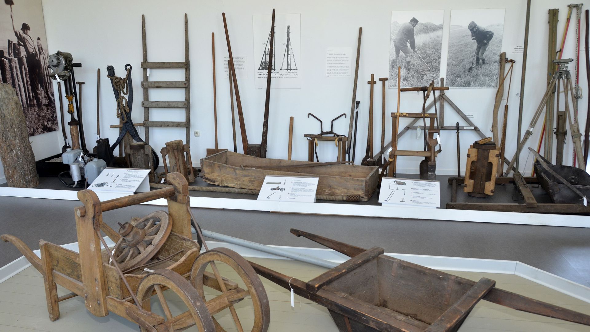 Ausstellungsraum des Deichmuseums Land Wursten in Dorum. Im Vordergrund befinden sich historische Holzwagen. Im Hintergrund sind verschiedene Werkzeuge und Geräte aus Holz und Metall ausgestellt, die für den Deichbau verwendet wurden. Tafeln und Bilder erläutern die historischen Arbeitsmethoden.