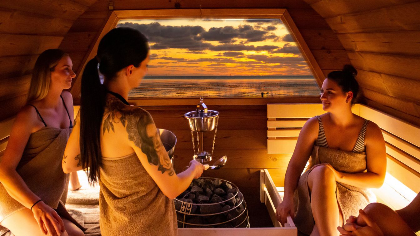 Es sitzen vier Frauen in der Fass-Sauna mit Handtuch auf einer Holzbank, sie unterhalten sich und im Hintergrund ist der Sonnenuntergang zu sehen.
