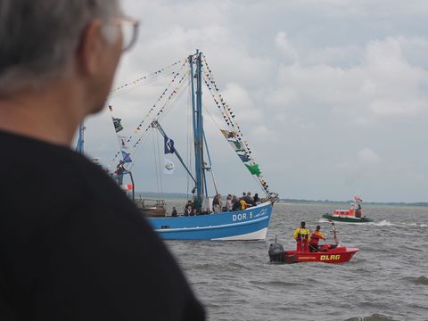 Beim Kutterkorso während der Dorumer Hafentage fährt ein blauer Fischkutter mit der Aufschrift ‚DOR 5‘ über das Wasser. Das Schiff ist mit bunten Flaggen und Wimpeln geschmückt und trägt mehrere Personen an Bord. Rechts daneben befindet sich ein rotes Rettungsboot der DLRG mit drei Personen in Rettungswesten sowie ein weiteres grün-weißes Boot im Hintergrund. Der Himmel ist bewölkt.