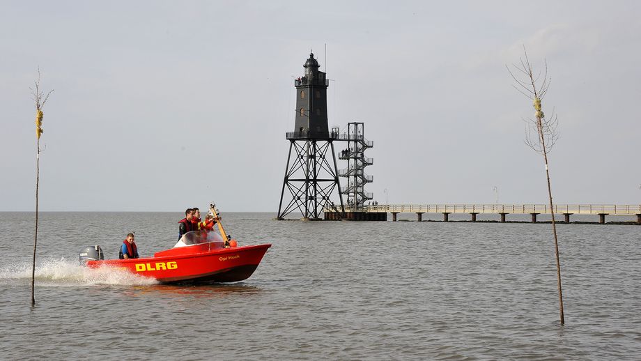 Ein rotes Motorboot der DLRG-Ortsgruppe Dorum fährt über das Wasser. Auf der Seite des Bootes steht gut sichtbar ‚DLRG‘. An Bord befinden sich mehrere Personen mit Rettungswesten. Im Hintergrund erhebt sich der Leuchtturm Obereversand. Im Vordergrund ragen zwei Pricken aus dem Wasser. 